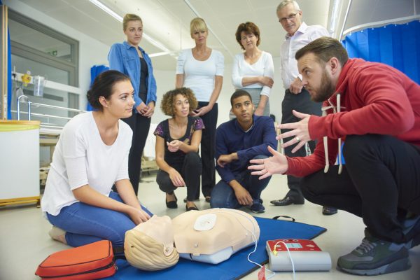 a mixed age group listen to their tutor as he shows the procedure involved to resuscitate using a defibrillator .