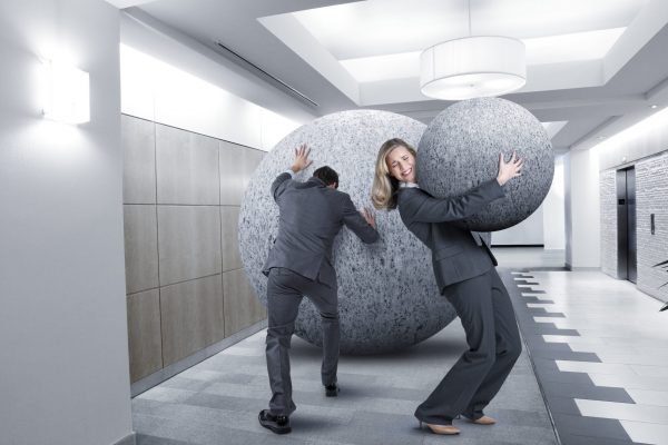 A businessman pushes a and a businesswoman struggles to carry a large stone in an office lobby.